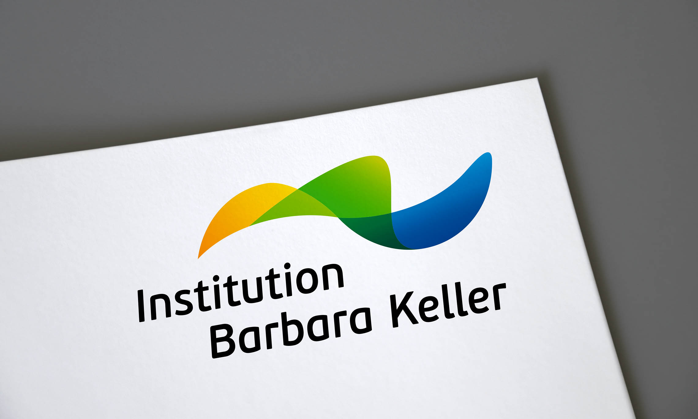 Das Logo der Institution Barbara Keller besteht aus einem gelben, grünen und blauen Schwungelement. Gelb steht für den Bereich "arbeiten", grün für den Bereich "lernen" und blau für den Bereich "wohnen". Unter dem Signet steht Institution Barbara Keller in einer kursiven Schrift.