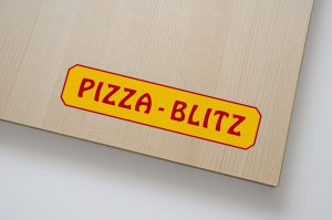 Das neue Logo von Pizza-Blitz auf einem Holzbrett. Es zeigt einen roten Schriftzug auf einer gelben Fläche. Die Fläche hat einen roten Rahmen und ist an den Ecken abgeschrägt.